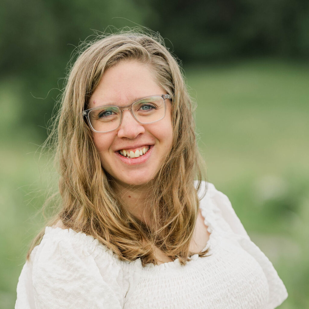 A photo of Danelle Lundstrom wearing a white dress with a blurred green natural background.